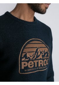 Marineblau strukturierter Pullover mit einem braunen gestickten Berg-Logo, auf dem "PETROL" sowie horizontale Linien darüber und darunter zu sehen sind.