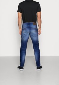 Blå denimjeans med slim fit, med en märkt läderpatch på bakstycket, kontrasterande sömmar och två bakfickor. Svart t-shirt och skor.