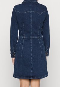 Kobieta ubrana w dopasowaną granatową sukienkę dżinsową z długimi rękawami i zapiętymi mankietami, pokazująca widok z tyłu z widocznymi detalami szwów.