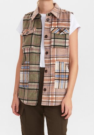 Mouwloos geruit overgooier met knopen, voorzien van twee borstzakken en twee onderste zakken, gedragen over een wit shirt met korte mouwen en een bruine broek.