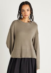 Oliwkowy sweter z dzianiny z prążkowanym dołem i szerokimi, opadającymi rękawami. Miękka tekstura, luźny krój. Nosić na ciemnej spódnicy.