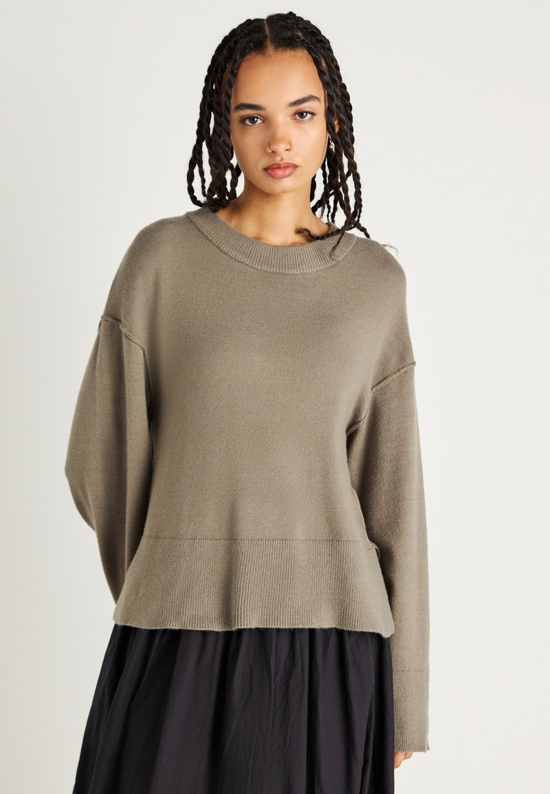 Oliwkowy sweter z dzianiny z prążkowanym dołem i szerokimi, opadającymi rękawami. Miękka tekstura, luźny krój. Nosić na ciemnej spódnicy.