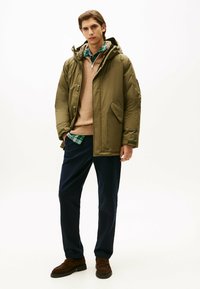 Doudoune vert olive avec capuche, superposée sur un pull beige et une chemise à carreaux. Pantalon bleu marine foncé et chaussures marron complètent la tenue.