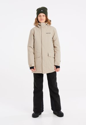 Giacca impermeabile beige con colletto alto, tasche laterali e logo sul petto, abbinata a pantaloni neri e un berretto verde scuro.