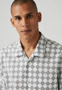 Camisa de manga corta en blanco con un patrón geométrico negro, con cuello y botones en el frente, hecha de una tela suave.