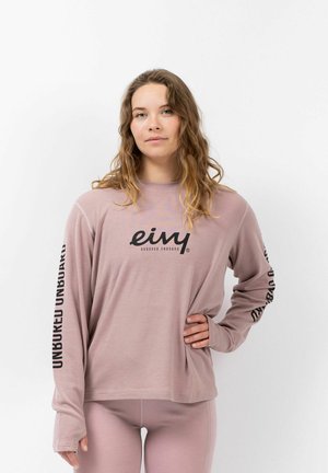Helles rosa Langarmhemd mit gerippter Textur, ausgestattet mit einem schwarzen Logo auf der Brust und schwarzem Text entlang der Ärmel. Kombiniert mit passender Hose.