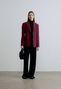 Bordeauxblazer met getailleerde pasvorm, zwarte coltrui, zwarte broek met brede pijpen en een kleine zwarte handtas. Soepele stof met een gestructureerd ontwerp.