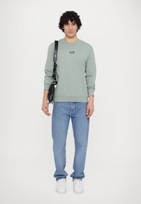 Felpa verde chiaro con collo rotondo e logo stampato, abbinata a jeans di denim blu chiaro e sneakers bianche. Mostrata una borsa a tracolla.