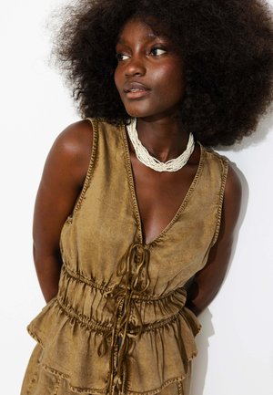 Femme aux cheveux bouclés naturels portant une robe marron sans manches et un collier de perles blanc, regardant sur le côté sur fond blanc.