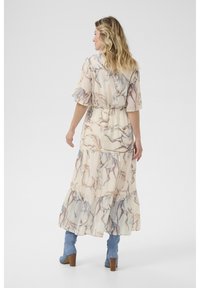 Maxikleid aus hellbeigem Stoff mit Pferdedruck, kurzen Glockenärmeln, taillierter Taille und gestuften Rüschen am Saum. Kombiniert mit blauen Stiefeletten mit Absatz.