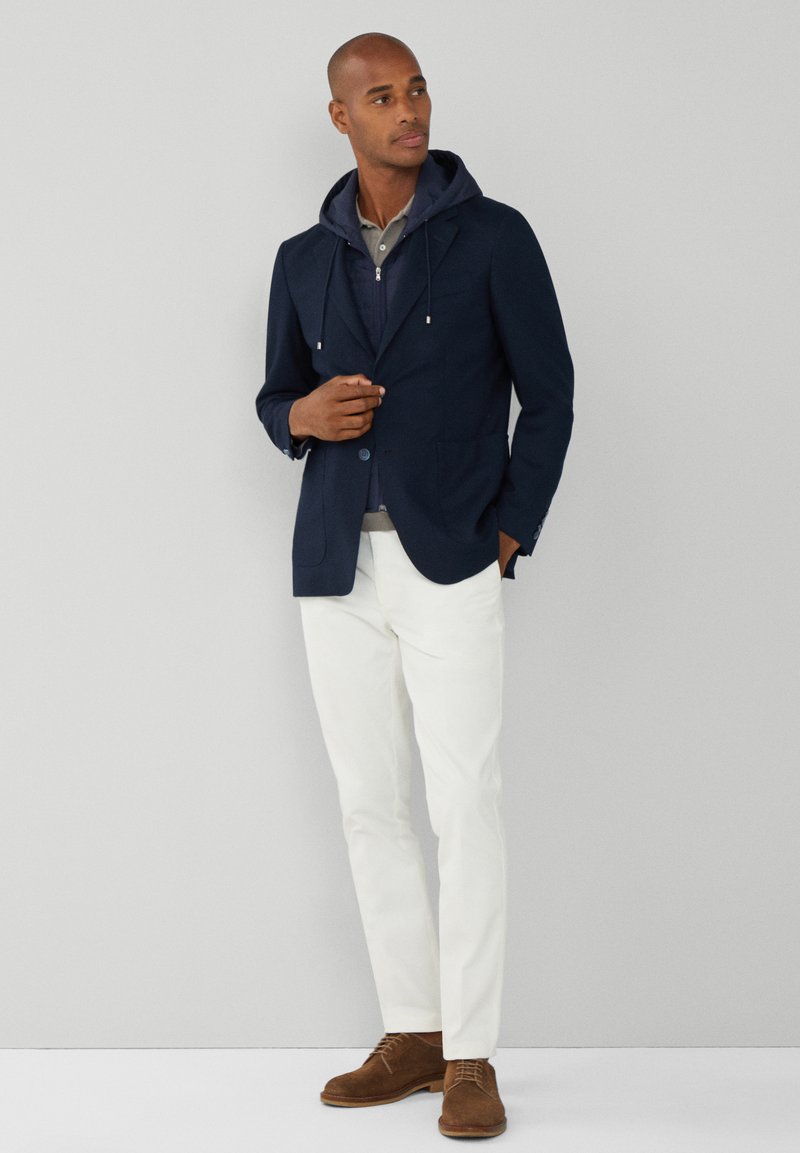 Blazer navy con diseño de doble botonadura, pantalones blancos y zapatos de ante marrón. Superpuesto con una sudadera gris con capucha y una camisa con cuello debajo.