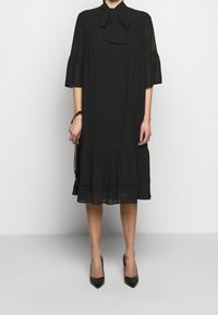 Robe noire avec un col montant, un détail en nœud, des manches évasées et un ourlet froncé, fabriquée en tissu léger. Associée à des talons noirs.