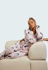 Rosa pyjamasset med puffin-mönster, bestående av en knäppningstopp och byxor. Mjuk tygtextur, avslappnad passform och en böjd beige soffa i bakgrunden.