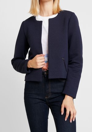 Kvinde iført en marineblå struktureret lynlåstrøje over en hvid grafisk t-shirt og mørkeblå jeans, poserer med den ene hånd, der holder jakken åben.