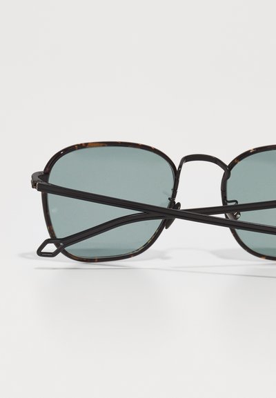 Ochelari de soare cu ramă din material cu model de broască țestoasă, accente negre și lentile de culoare verde. Prezintă o formă pătrată și brațe negre subțiri, intersectate.