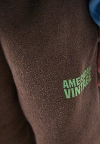 Pruun kangas, mille paremas alanurgas on helesinise varrukaga käe poolt osaliselt kaetud helerohelise tekstiga "AMERICAN VINTAGE".