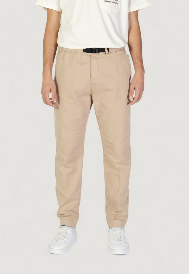 Pantaloni in cotone beige con vestibilità rilassata, dotati di una cintura elasticizzata e una cintura nera, abbinati a sneakers bianche.