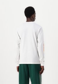 White long-sleeve shirt featuring a crew neckline and multicolor graphic on the left sleeve, paired with green pleated shorts.