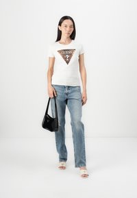 Hvid bomulds T-shirt med et trekantet brunt mønsterdesign og "GUESS" tekst, parret med lyseblå jeans og en sort skuldertaske.