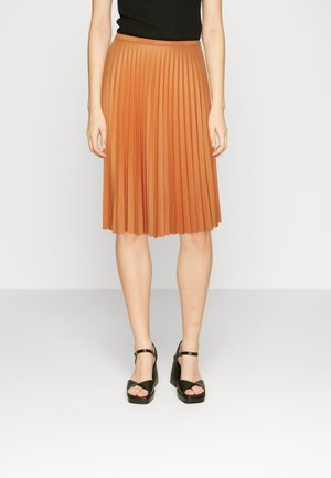 Mujer vestida con una falda plisada naranja hasta la rodilla y una blusa sin mangas negra, combinadas con tacones gruesos negros con correa en el tobillo, sobre un fondo blanco.