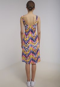 Femme avec un chignon portant une robe sans manches à motif zigzag coloré et des baskets blanches, tournée vers le dos contre un fond uni.