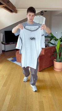 Lys grå oversize t-shirt med et sort logo på forsiden, holdt af en person. Indendørs miljø med trægulv og planter i baggrunden.
