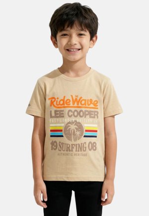 Garçon souriant portant un t-shirt graphique beige « Ride The Wave Lee Cooper » avec motif surf, arbre de palmier et rayures colorées, debout devant un fond uni.