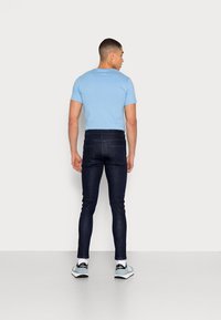 T-shirt azul claro de manga curta e jeans skinny azul escuro com uma textura suave, combinados com ténis cinza e brancos. Vista traseira.