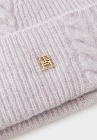 Lilla strikket hue lavet af blødt garn, med et struktureret ribmønster og en gylden logoaccent på den foldede kant.