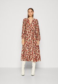Abito lungo beige con un vivace motivo animale rosso e viola. Presenta una scollatura a V, maniche lunghe e una vita cintrata. Indossato con stivali bianchi al ginocchio.