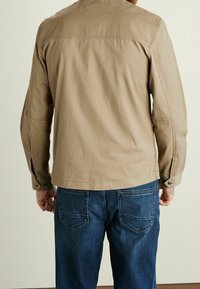 Light beige jacket with a relaxed fit, featuring buttoned cuffs and seams, paired with dark blue denim jeans. Smooth fabric texture.