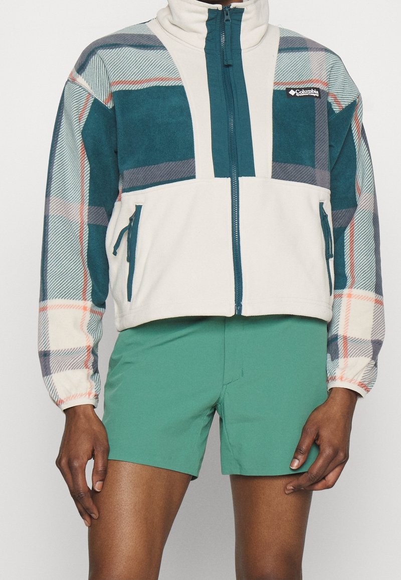 Bluza polarowa z beżowym korpusem i rękawami w kratę w kolorach morskim, szarym i czerwonym; posiada wysoki kołnierz i kieszenie na zamek. Połączona z zielonymi szortami.