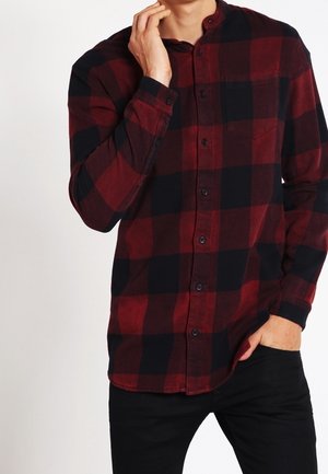 Mand iført en rød og sort ternet langærmet skjorte med knapper, med den ene hånd ved ansigtet og den anden i lommen.