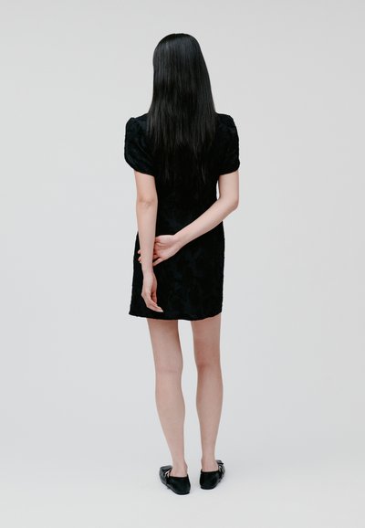 Black dress with textured fabric, short puff sleeves, and a fitted silhouette, paired with black flat shoes. Model is viewed from the back.