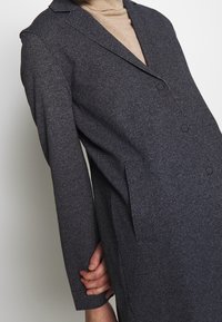 Harmaa, teksturoitu villasekoite takki, jossa on tuplapainike kiinnitys, vaaleat läpät ja sivutaskut. Kaupunkityyli, jossa on istuva leikkaus.