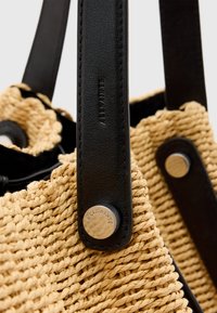Borsa tote in paglia intrecciata con manici in pelle nera, accent metallico rotondo e superficie testurizzata. Presenta un colore beige naturale e una costruzione resistente.