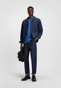 Denimjakke, blå skjorte, mørke jeans og sorte støvler. Modellen holder en sort taske. Alle stykker har en moderne pasform og minimalistisk design.