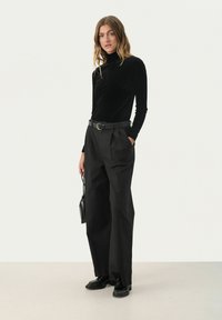 Schwarzer Samtrollkragenpullover kombiniert mit hochtaillierten schwarzen Hosen. Die Hosen verfügen über scharfe Falten und ein weites Bein-Design. Zubehör umfasst eine schwarze Handtasche.