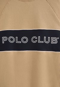 Felpa beige con una banda orizzontale blu navy che presenta il testo "POLO CLUB" in bianco. Realizzata in morbido tessuto con colletto e polsini a costine.
