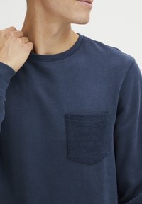 Maglietta a maniche lunghe blu navy realizzata in tessuto morbido, con una tasca a toppa texturizzata sul lato sinistro del petto e un colletto rotondo.
