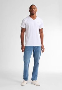 Tricou alb din bumbac cu guler în V și mâneci scurte, asortat cu blugi bleu deschis și adidași bej. Design simplu cu un mic logo pe piept.