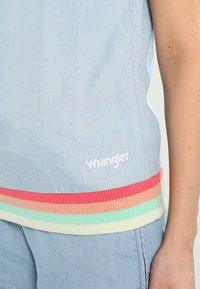 Top azzurro chiaro senza maniche con orlo a righe multicolori a costine. Presenta il logo "Wrangler" in bianco sul davanti, tessuto di consistenza liscia.