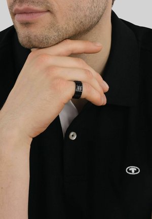Mann mit leichtem Bartschatten, der ein schwarzes Hemd mit Kragen und einen schwarzen segmentierten Ring am Zeigefinger trägt, der nahe seinem Kinn ruht.