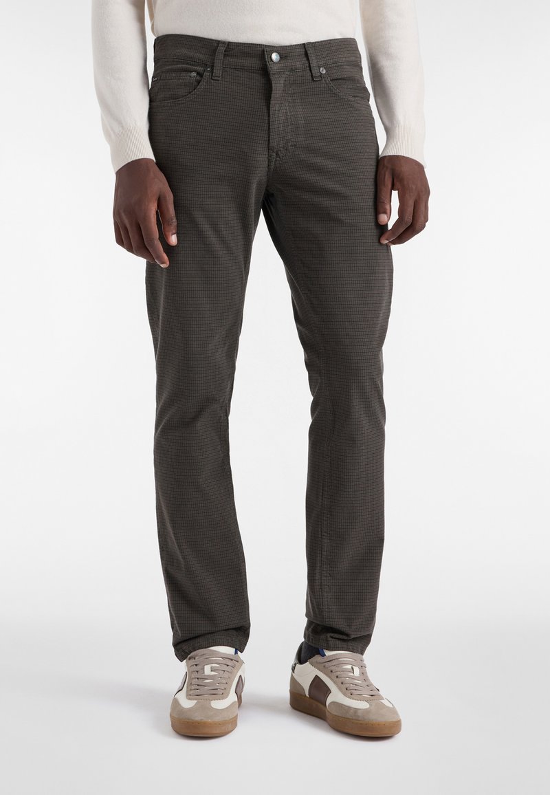 Moški model nosi temno karirane slim-fit hlače in bele superge z rjavimi akcenti, stoji ob navadni beli ozadju.