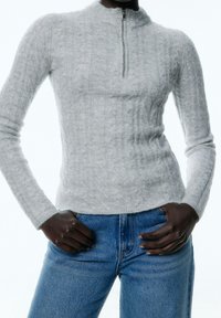 Ljust grå zip-up-tröja med ett texturerat kabelstickat mönster och hög krage, parat med blå denimjeans.