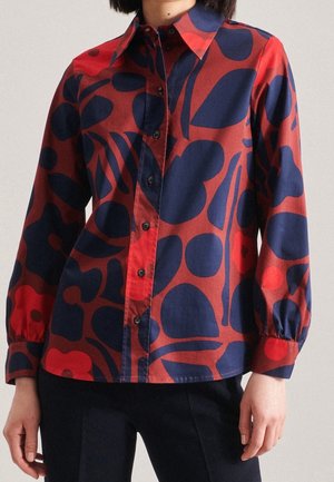 Donna che indossa una camicia rossa con bottoni, decorata con grandi motivi floreali astratti blu navy e rossi e maniche lunghe a sbuffo.