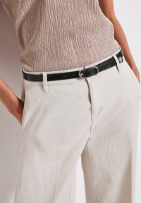 Valkoiset leveälahkeiset housut, joissa on etutaskut, ja teksturoitu taupe-välinen paita, yhdistettynä mustaan nahkavyöhön, jossa on hopeanvärinen soikea solki.