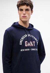 Granatowa bluza z kapturem z przednią kieszenią typu kangurek, kapturem z suwakiem oraz białym/czerwonym napisem "American Sportswear GANT New Haven - Connecticut."