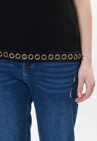 Top nero con occhielli decorativi lungo una fascia testurizzata; abbinato a jeans in denim blu strappati. Il tessuto appare liscio.
