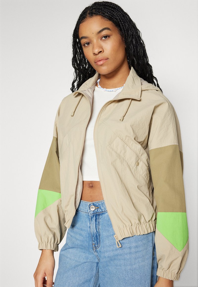 Chaqueta ligera beige con detalles en verde y verde oliva, puños elásticos y cierre de cremallera. Ajuste casual sobre una blusa blanca y jeans de mezclilla.